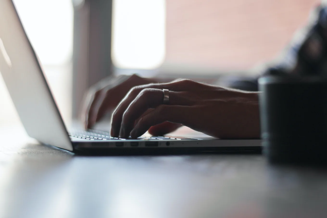 Hands typing on a laptop keyboard, blurred cup.